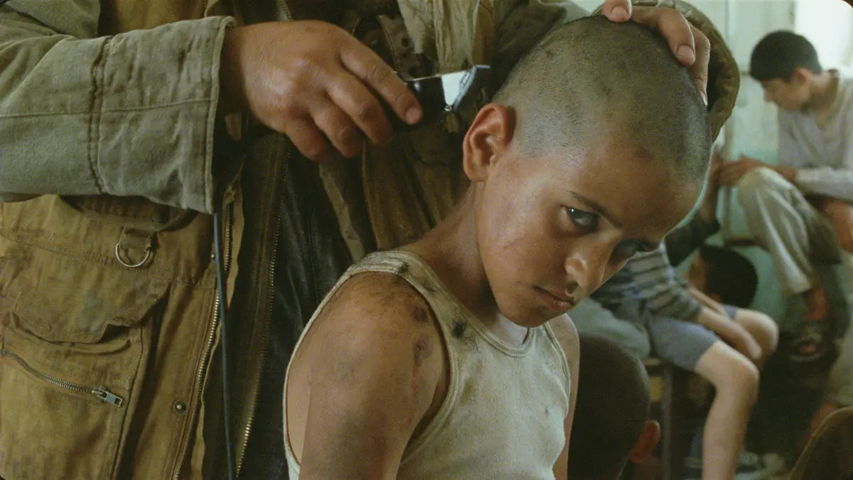 menino olha com expressão séria enquanto um homem raspa a cabeça dele em cena do filme Incêndios de 2010