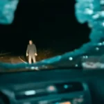 vidro dianteiro de carro quebrado com homem iluminado na estrada em cena do filme de terror Passageiro do Mal de 2026