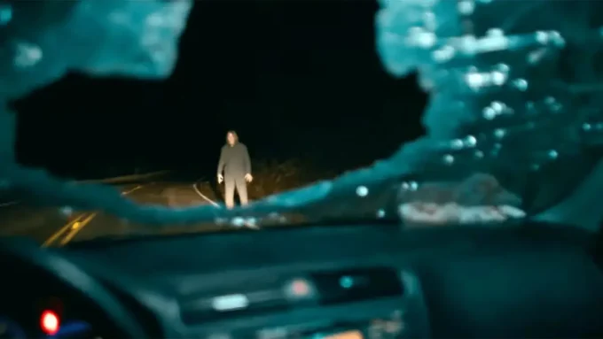 vidro dianteiro de carro quebrado com homem iluminado na estrada em cena do filme de terror Passageiro do Mal de 2026